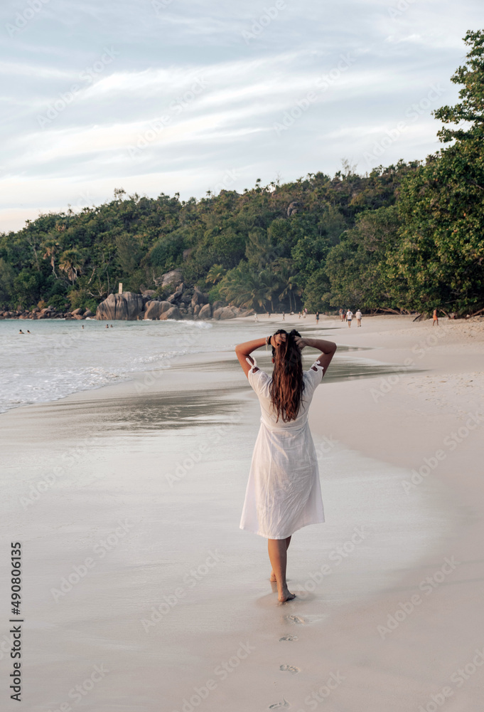 Sticker Vertical view of the Caucasian female walking on the sandy beach with white dress in Seychelles