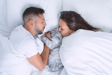 Young couple lying in the bed with adorable cat. Woman kissing cute cat. Love for cats or pets. Sleep with pets