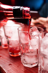 Red syrup is poured from a bottle into a large cocktail glass with ice cubes on the bar counter