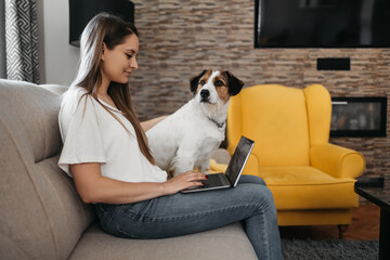 Young woman sitting on sofa with adorable adopted dog in her home living room and using laptop computer.