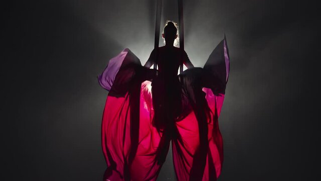 Silhouette of an aerial circus gymnast performing on aerial silk and waving canvas like wings. Beautiful circus show performed by young woman acrobat on black background with backlight. Slow motion.