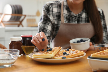 Woman decorating delicious Belgian waffles with blueberries at wooden table in kitchen, closeup