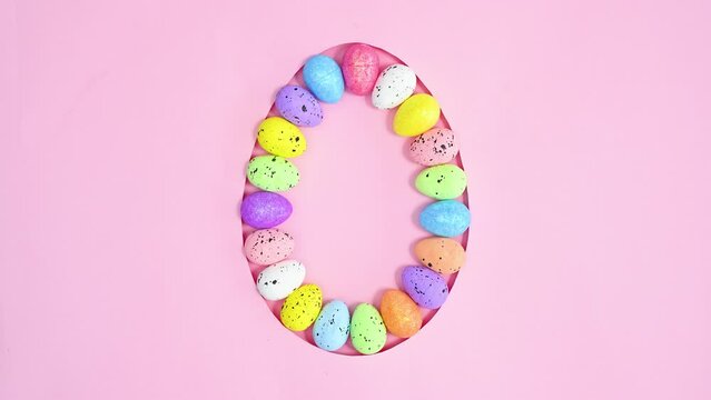 Colorful Easter eggs appear in egg shape frame on pastel pink background. Creative stop motion flat lay