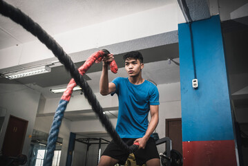 A fit young asian man working out vigorously with battle ropes. Alternating single arm waves. Whole...