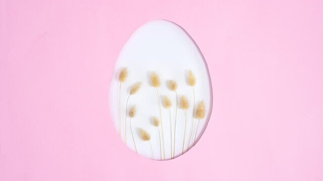 Dried trendy fashion plants appear in Easter egg shape on pastel pink background. Stop motion flat lay