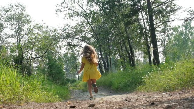 Happy little girl with blond long hair in a yellow dress and running  along path on meadow with sunset