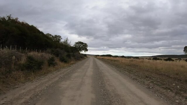 Countryside scene. Point of view of a car speeding across the dirt road in the farmland in a cloudy day. We can see the ranch forest and yellow grassland in autumn.