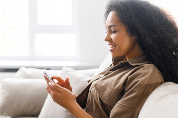 Side view at young multiracial woman using trendy smartphone sitting on the sofa at home, ethnic...
