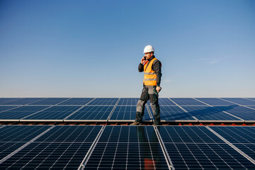 A repairman talking on the phone while standing on rooftop surrounded by solar panels.