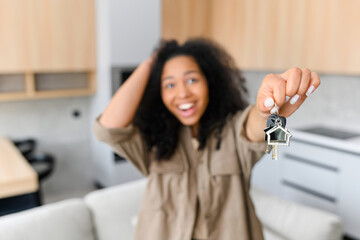 Joyful multiracial woman holding keys with trinket in shape of little cute house and showing it to...