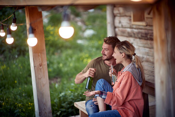 Young cheerful couple having a conversation and drinking beer in front of wooden cottage on the terrace.  Summertime garden celebration and fun. Friends, togetherness, fun, joy, celebration concept.