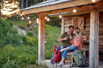 A young couple having chatting and having a drink at the cottage porch in the forest. Vacation,...