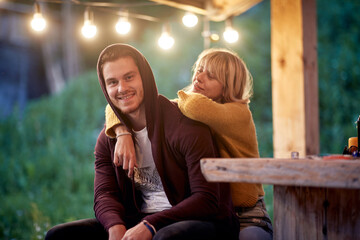 Young attractive cheerful couple sitting in front of wooden cottage. Woman leaning on to her boyfriend and hugging him. Beuatiful moments. Vacation, holiday, togetherness concept.