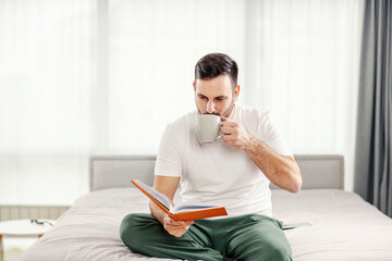 A morning man drinking his coffee and reading a book on the bed at his cozy apartment.