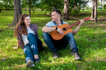Fototapeta premium Young couple on the grass in the park in the summer, a guy plays the guitar for a girl