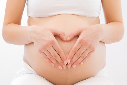 Heart shape created from young adult pregnant woman hands on big naked belly on white background. Pregnancy time. Baby expectation. Love, happiness and safety concept. Closeup. Front view.