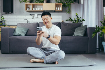 Obraz premium Male asian athlete at home using phone sitting on the floor during morning exercise, athlete in living room