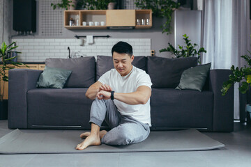 Asian at home smiling, doing fitness in the morning, in the living room, choosing a sports program on a fitness smart watch, a man leads an active lifestyle