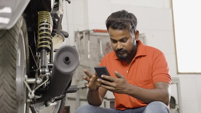 Mechanic using mobile phone in front of motorbike at garage - concept of using social media at work, taking break and seraching tools or repair parts online ofor shopping.