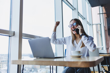 A young female manager in glasses works at a laptop and gesticulates emotionally with her hands while talking on the phone, she is sitting at her desk in a white shirt