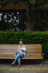 A young mother is breastfeeding a baby sitting on a bench in the park. Breast-feeding with milk. Mom in a striped jacket and jeans. A child in white pants and a hat.