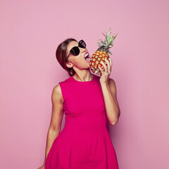 Fashion portrait of healthy woman biting pineapple over colorful pink background