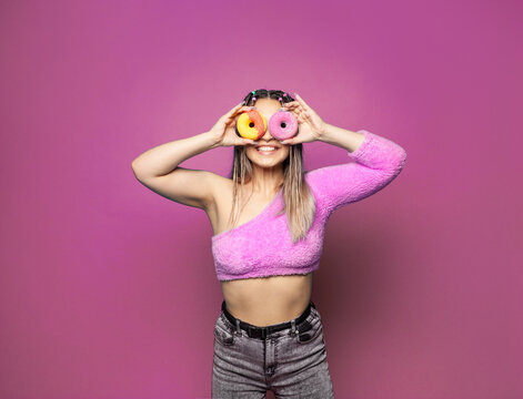 Beauty fashion model woman with colorful sweet donuts. Funny joyful stylish woman with sweets on pink banner background. Diet, dieting and party concept. Junk food, slimming, weight loss