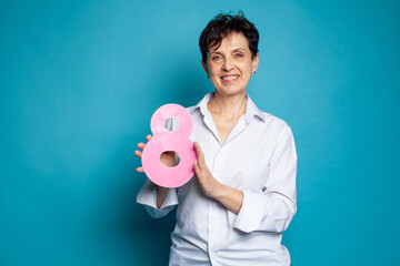 Portrait of happy mature model woman in white shirt holding pink eight 8 sign banner on colorful blue studio wall background