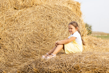 Portrait of happy little girl with eyes squinted from sun and long braid on head looking away sitting on haystacks. Time away from city in country field with tons of hayricks.