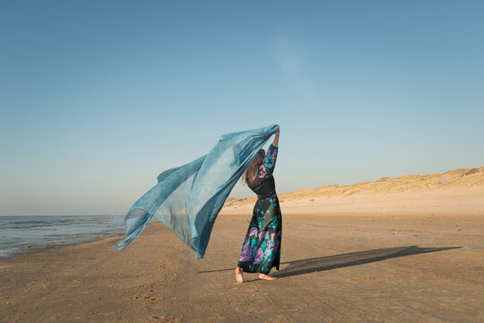woman on the beach in long floral dress dancing with blue sheet near ocean in sun