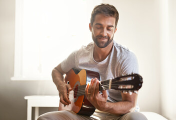Honing his musical talent. Shot of a handsome young man playing a guitar at home. - obrazy, fototapety, plakaty