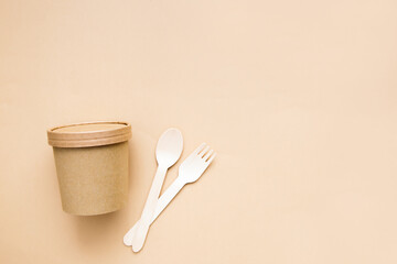 Top view of wooden fork, spoon and paper cup on a beige background. Natural eco friendly disposable tableware or creative design idea. Copy space.