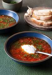 Homemade borscht with sour cream and herbs in blue plates on a green background with bread and garlic