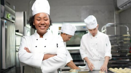 Selective focus of African female baker in a chef dress and hat, standing with arms folded and smiling at camera, with blurred colleagues kneading the dough in the background. Copy space on right side