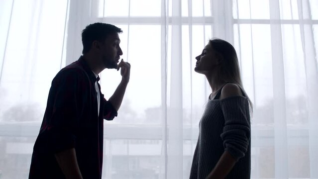 Married couple swears in living room background of curtains and windows, husband insults wife by pointing at her with finger, side view. Husband and wife quarrel at home. Concept of family problems
