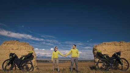 The man and woman travel on mixed terrain cycle bike touring with bikepacking. The two people journey with bicycle bags. Sport sportswear in green black colors. Mountain snow capped, stone arch.