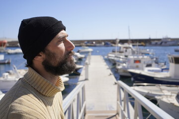 profile portrait of bearded man wearing turtleneck and hat looking at camera in a harbor © javiemebravo