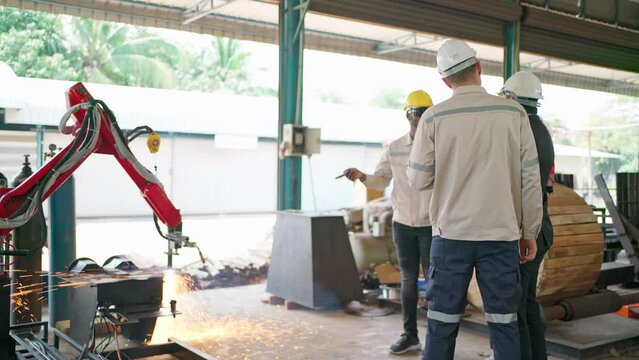 Engineer discuss and brainstorm in the automate robotic arm factory. Manager and team looking at robot arm operate. Welding tool weld metal. Bright sparks at production. Teamwork and technology