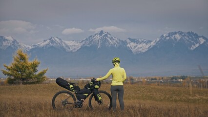 Obraz premium The woman travel on mixed terrain cycle touring with bikepacking. The traveler journey with bicycle bags. Sport tourism bikepacking, bike, sportswear in green black colors. Mountain snow capped.