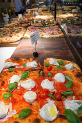 A close-up shot of a pizza in a shop with a tomato base, mozzarella cheese, cherry tomatoes, and basil as a garnish, with other pizzas in the background.  Image features selective focus. - obrazy, fototapety, plakaty