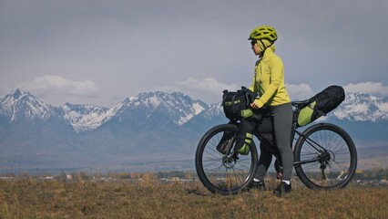 Naklejka premium The woman travel on mixed terrain cycle touring with bikepacking. The traveler journey with bicycle bags. Sport tourism bikepacking, bike, sportswear in green black colors. Mountain snow capped.