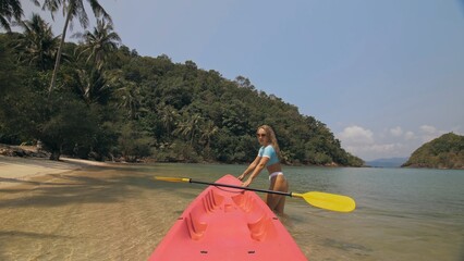 Young woman in sunglasses pulls pink plastic canoe with paddle to azure sea water from beach with...