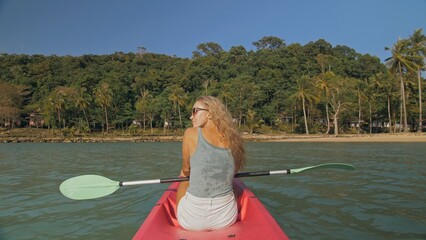Long haired blonde woman with sunglasses rows bright pink canoe along sea bay water to beach with growing palms. Traveling to tropical countries. Sports girl is sailing on kayak in ocean. - obrazy, fototapety, plakaty