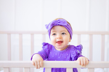 cute portrait of a baby girl in a crib holding on to the side in lilac clothes and with a bow on...
