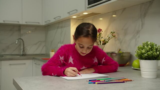 Lovely preteen girl in pink clothing developing inherent capacities, skills, creativity talents and hobby at home. Charming female kid drawing with colorful pencils in sketchbook indoors. Moving shot