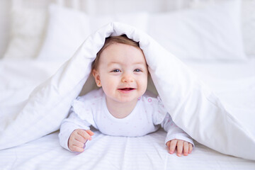 funny happy baby girl looks out from under the blanket on the bed in white clothes and smiles, a...