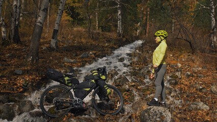 The woman travel on mixed terrain cycle touring with bikepacking. The traveler journey with bicycle bags. Sportswear in green black colors. The trip in magical autumn forest, river stream.