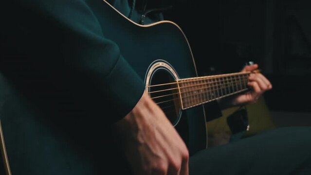 Man plays a pick on a modern guitar in the dark slow mo. A young musician plays the guitar in the living room at home. Relax with song and music. He has fun playing the acoustic guitar. Close-up.