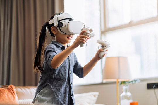 exiting asian child female enjoy metaverse gaming with wearable vr headset with control handle playing sport gaming online in living room at home,home technology female daughter using vr technology