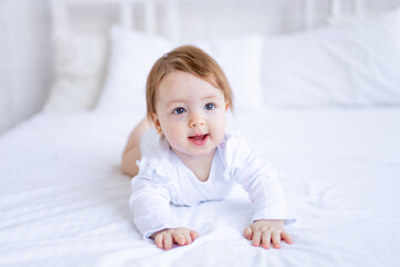Fototapeta premium funny baby girl of six months on a bed in white clothes smiling, a small child on a cotton bed at home woke up in the morning after sleeping, the concept of children's goods